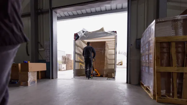 Man uses a pallet jack to move a stack of cardboard boxes while facing a trailer at a warehouse.