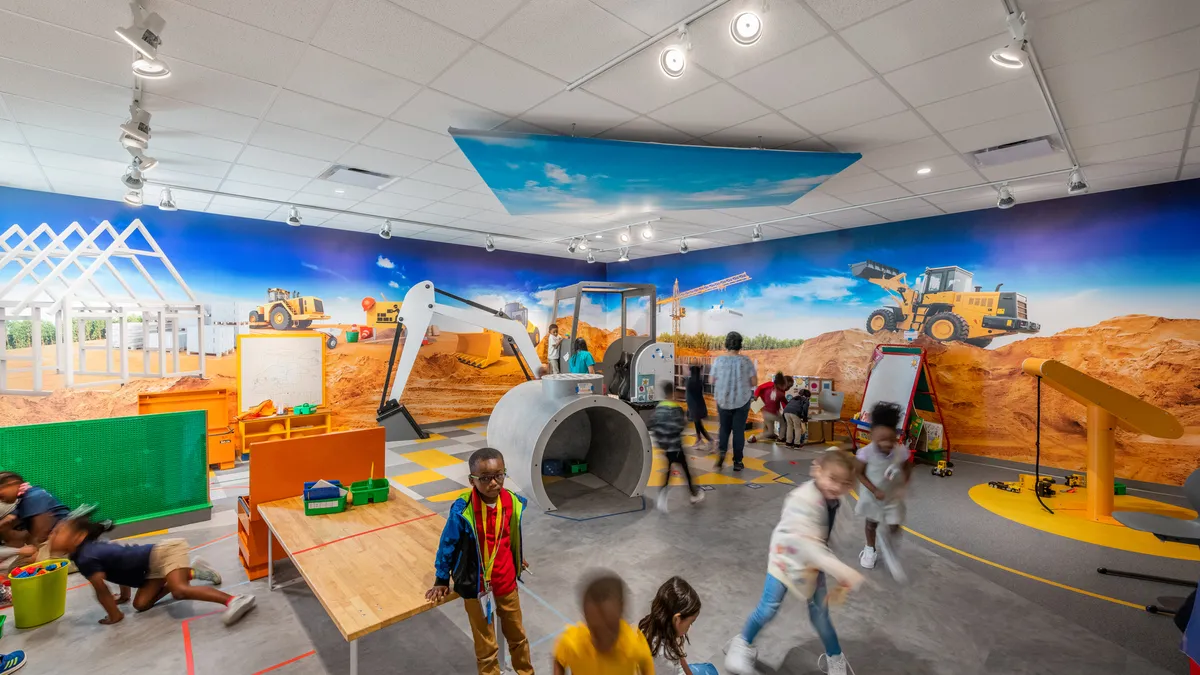 A group of young students are moving around an indoor play space that has a construction theme. There are kid-sized versions of a crane and backhoe. A colorful mural is on the wall.