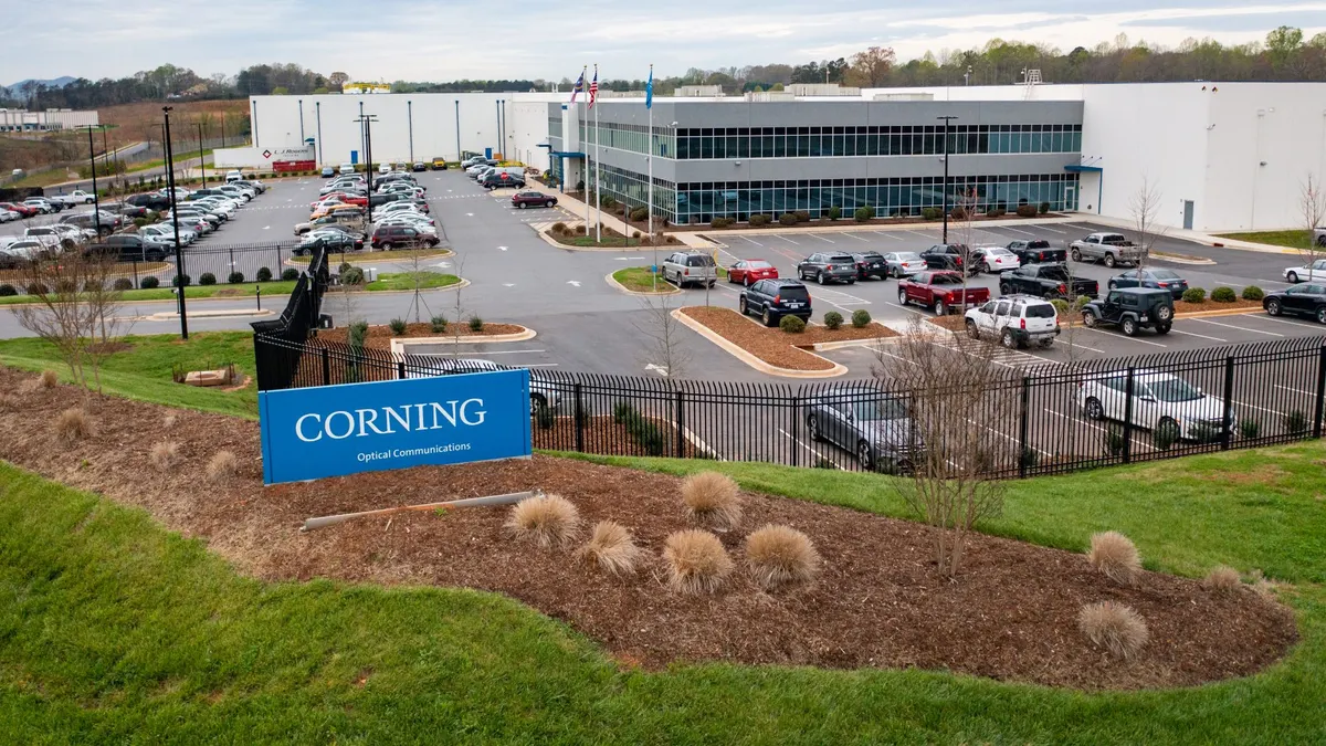 A blue and white Corning sign on a hill, with a large facility and a parking lot in the background.