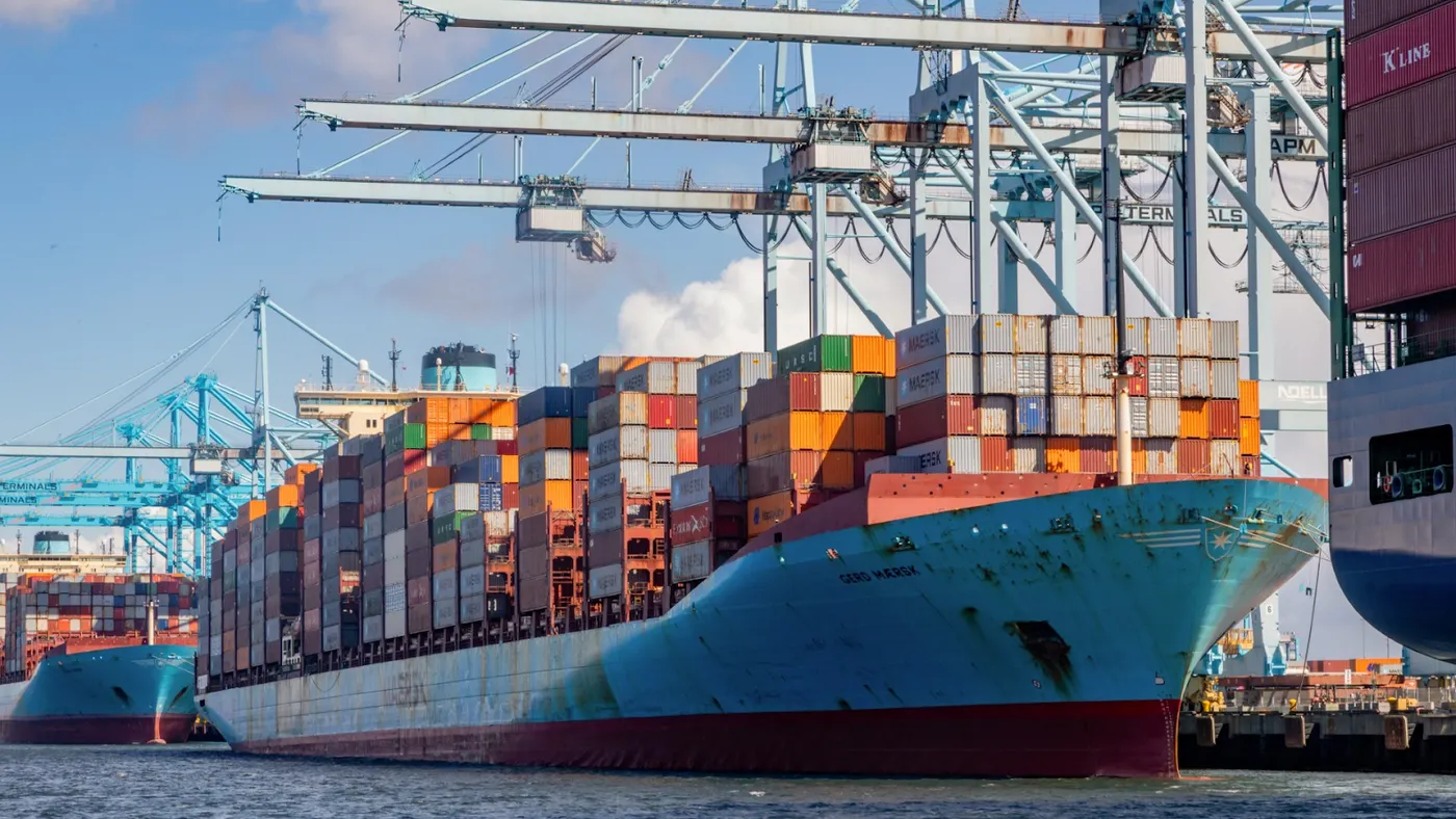 Dozens of multi-colored containers are stacked on a blue cargo ship docked at a port under cranes. The cargo ship is between two other vessels.
