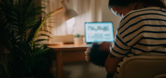 Woman breastfeeds baby while in front of laptop with lamp on.