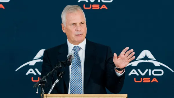 A person with short gray hair in a dark blue suit standing behind a light tanned podium.