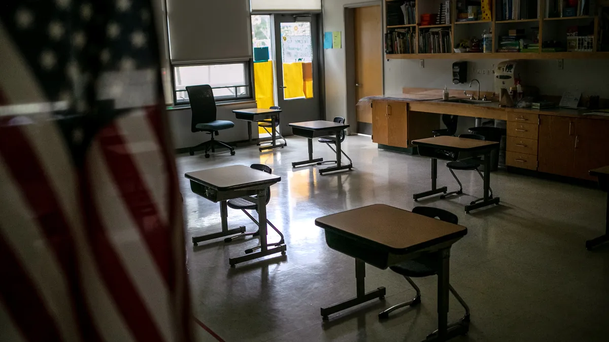 An American flag hangs in the foreground of an empty classroom with desks.