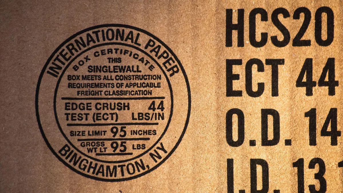 Close-up view of a printed seal on a brown cardboard box. The black circular stamp includes product details and is from "INTERNATIONAL PAPER" located in "BINGHAMTON, NY." Additional printed text is visible on the right-hand side in large black font.