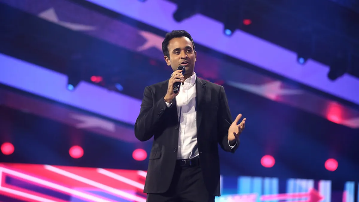 Vivek Ramaswamy stands with a microphone with pink and blue lights flashing behind him.