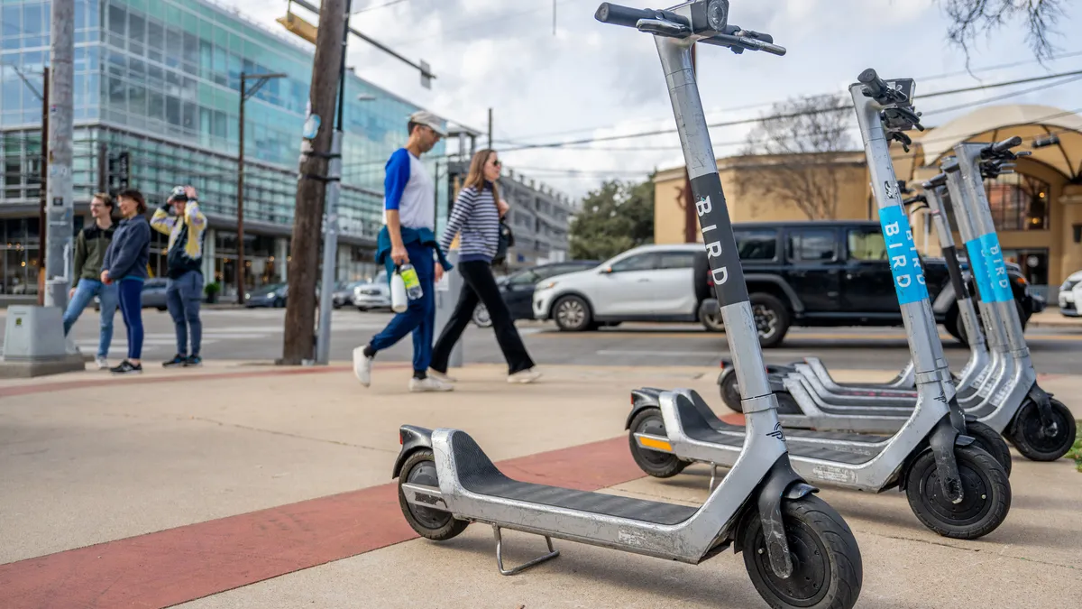 Several electric scooters on the sidewalk with people walking behind them.