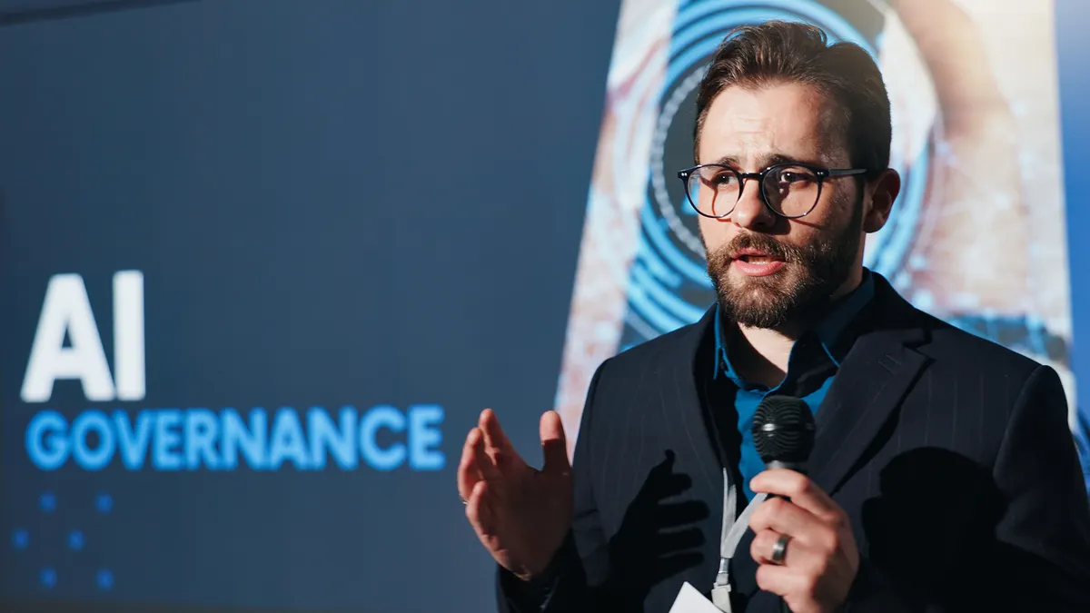 A man in glasses speaks into a microphone at an AI Governance conference. He gestures with his left hand, conveying a serious, engaged tone.