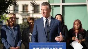 A person in a suit and tie stands behind a microphone at a podium. Behind him, several people watch.