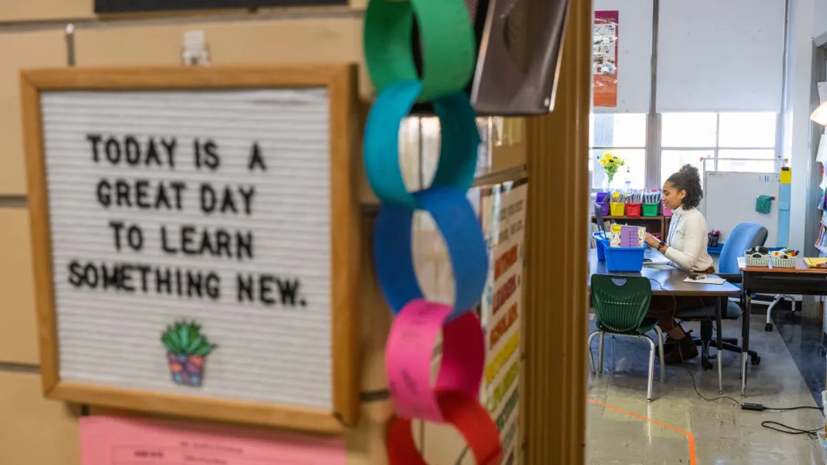 A person is seen sitting at a table in a classroom. The photo is taken from the door of the classroom and near the door is a sign reading "Today is a Great Day To Learn Something New"