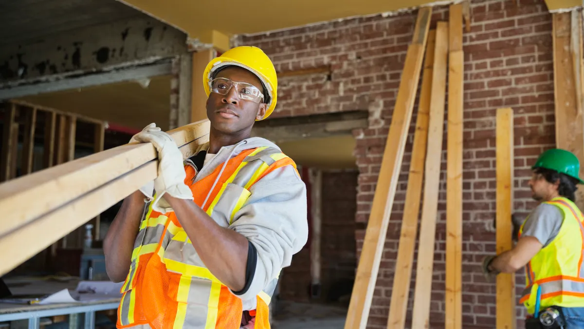 A worker in a hard hat and vest carries lumber at a jobsite.