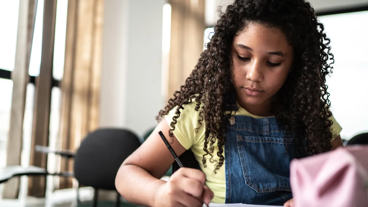 A girl student in a yellow t-shirt and overalls stares down at paper, pen in hand, writing notes in a notebook while silently contemplating classroom lessons.