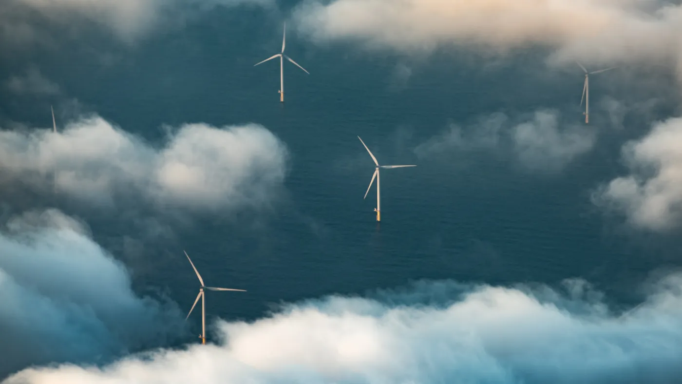 An overhead shot of the ocean shows overhead wind turbines, partially obscured by clouds.