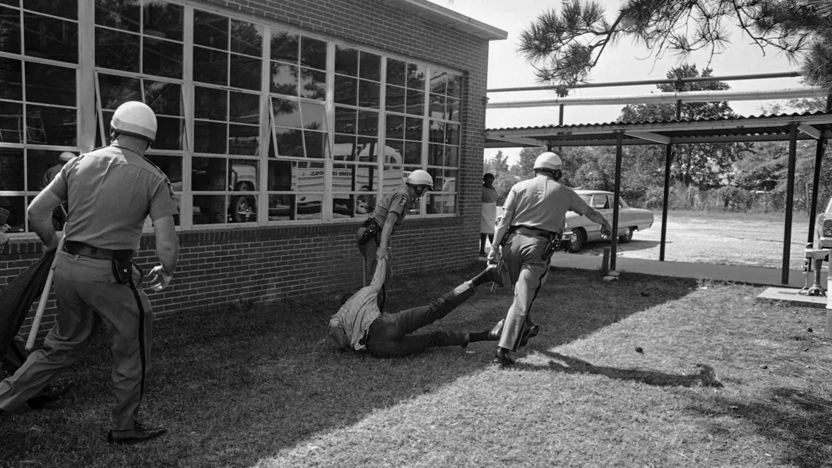 Georgia State Police are carrying away a Black demonstrator by their arm and leg outside a Black school in Georgia following a protest against segregated schools.