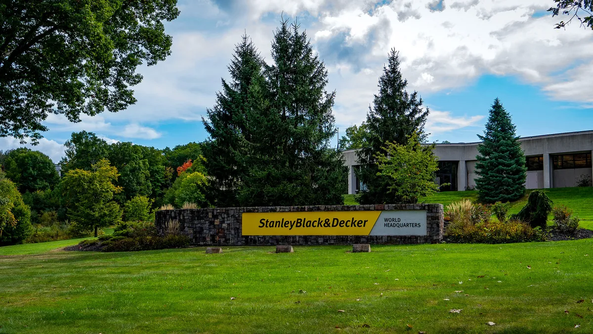 Black and yellow Stanley Black & Decker sign on green grass, and trees and a white building in the background on a sunny day.