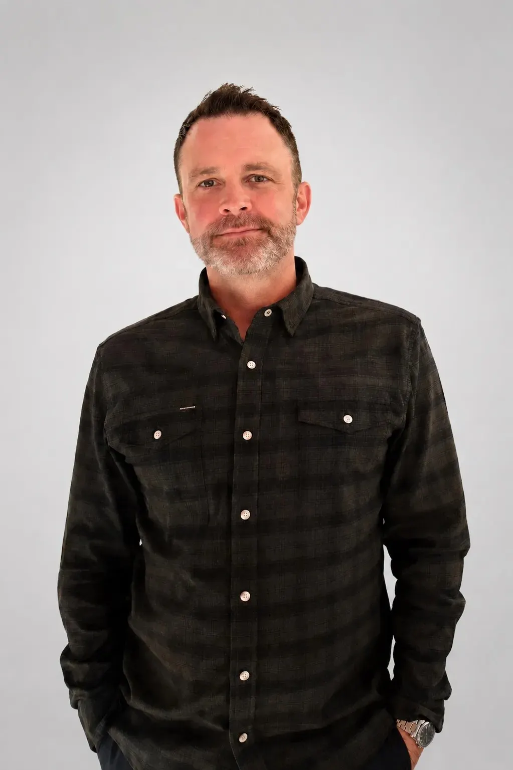 A white man in a dark shirt with stubble and a slight smile.