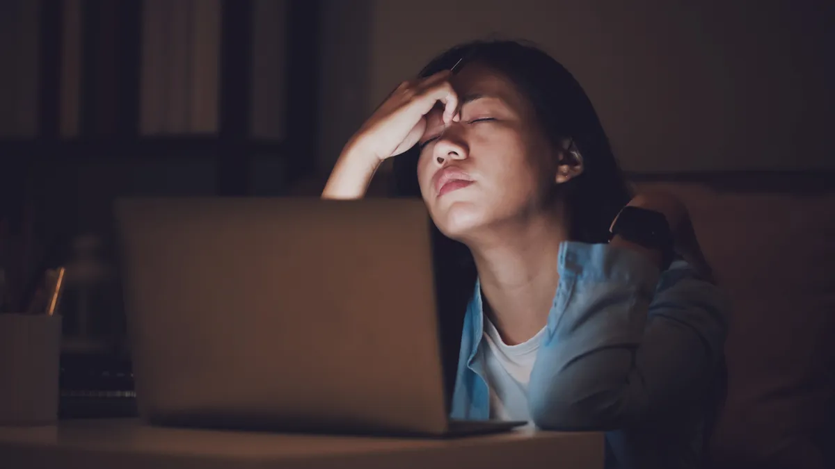 A person sits at a laptop in the dark, looking exhausted.