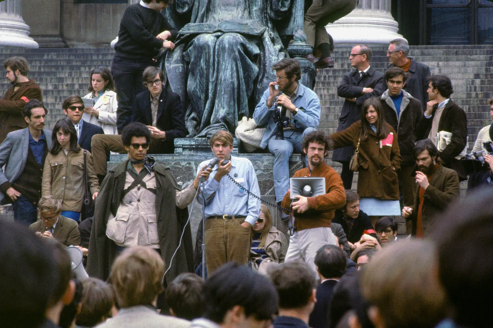 A person speaks into a microphone while addressing a crowd of students.