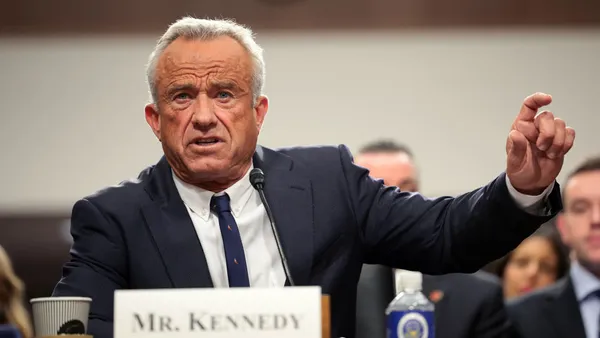 Robert F. Kennedy Jr. sitting with his name printed in front of a microphone in front of him with his arm extended besides him while speaking in a room with government officials in the background.