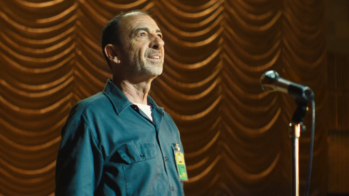 A janitor wearing a blue jumpsuit stands in the spotlight on a theater stage in Amazon's 2024 holiday marketing campaign