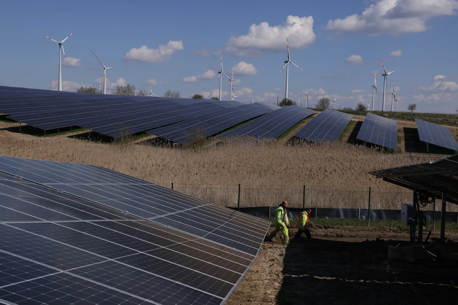 Workers walk among solar panels at the construction site of a new solar energy park as wind turbines spin behind on April 06, 2023 near Prenzlau, Germany.