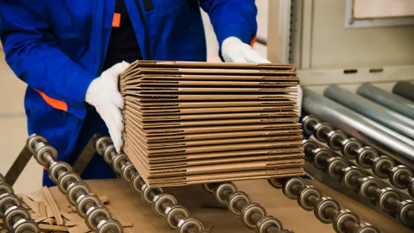 A person's hands are holding a stack of flat corrugated boxes next to a manufacturing production line.