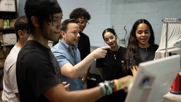A principal is pictured standing among students, speaking to them as they use an embroidery machine in a commercial art course.