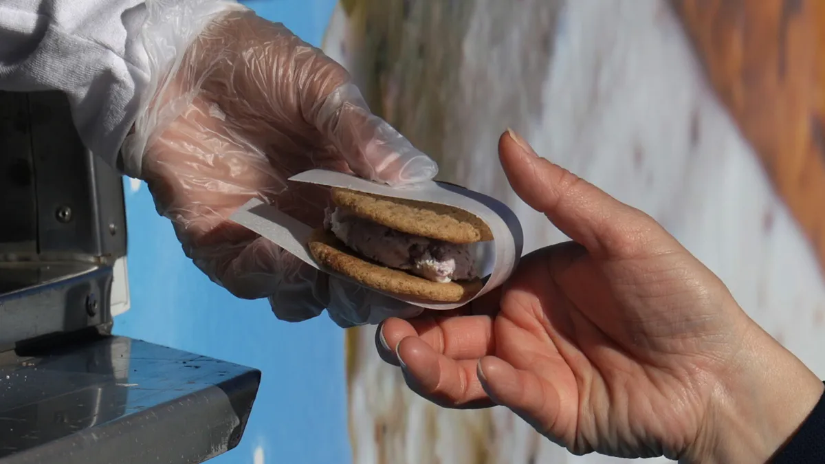 A hand wearing a safety glove gives a paper-wrapped ice cream sandwich with round cookies and pink ice cream to another hand.