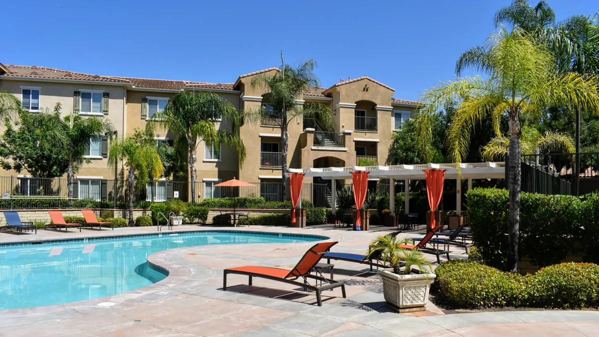 Tan, three-story apartment building with palm trees and a pool in the foreground.