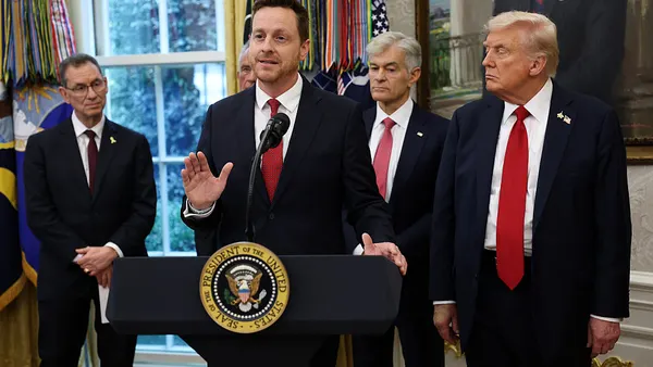 A pan wearing a black suit stands behind a podium flanked by other officials