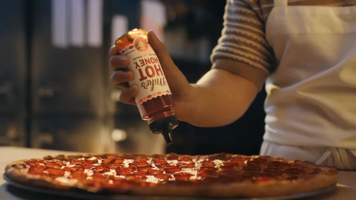 Mike's Hot Honey is drizzled onto a pepperoni pizza from a bottle with a white label by someone wearing a stripped brown polo shirt and white apron.