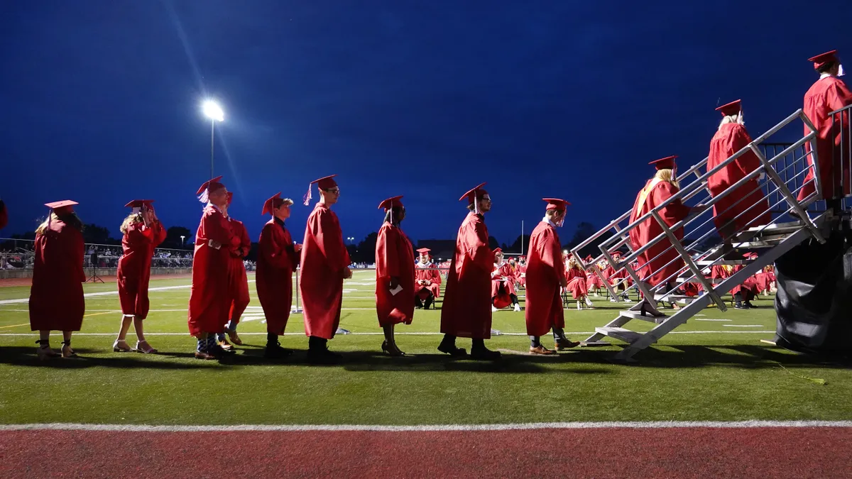 A line of students in red gowns stand on a football field, those on the right beginning to climb stairs to a stage.