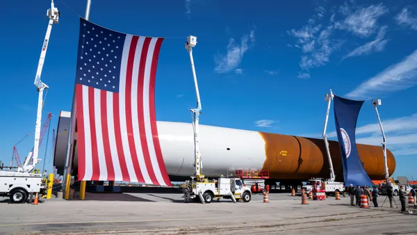 The United States flag and the Virginia state flag are shown in front of monopile foundations being delivered to a port in Virginia.