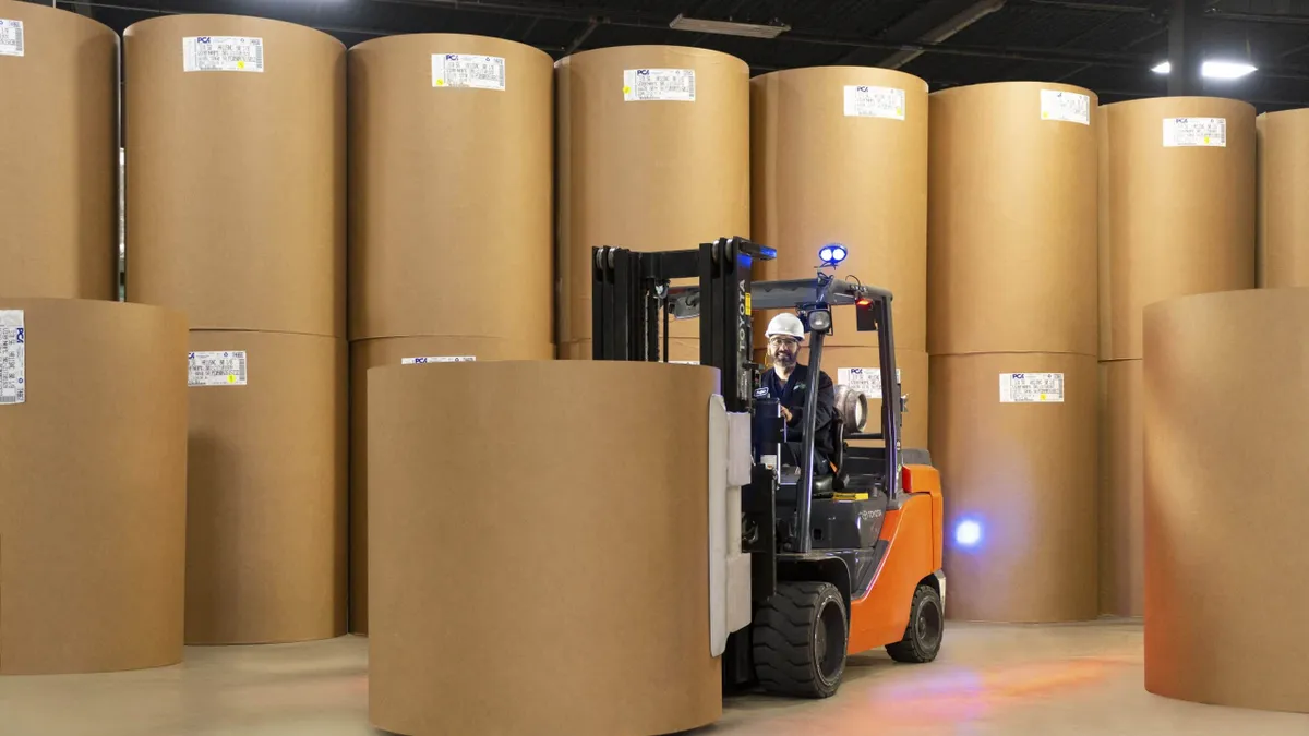 A worker operates a forklift in an industrial warehouse, surrounded by large rolls of a fiber product.