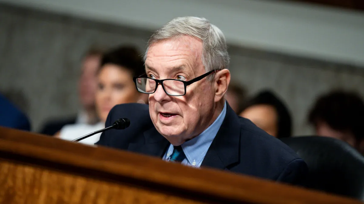 Man in glasses speaks at microphone seated on dias behind a wood railing.