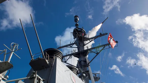A trail of smoke emitting from a missile in the blue sky as it leaves a gray ship.