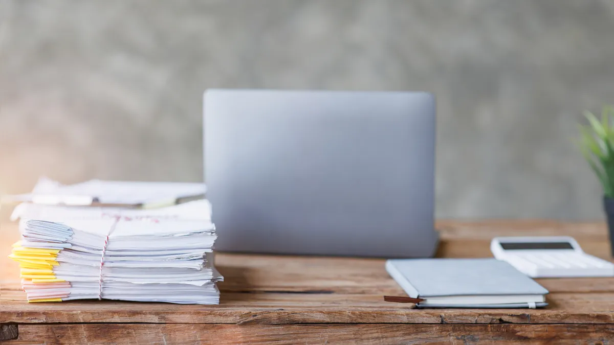 A laptop sits open on a table with the back facing the camera. Also on the desk is a stack of papers, a calculator, a plant and a journal.