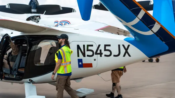 A person wearing a visibility vest walks next to a small white and blue aircraft as another person holds onto the front door.