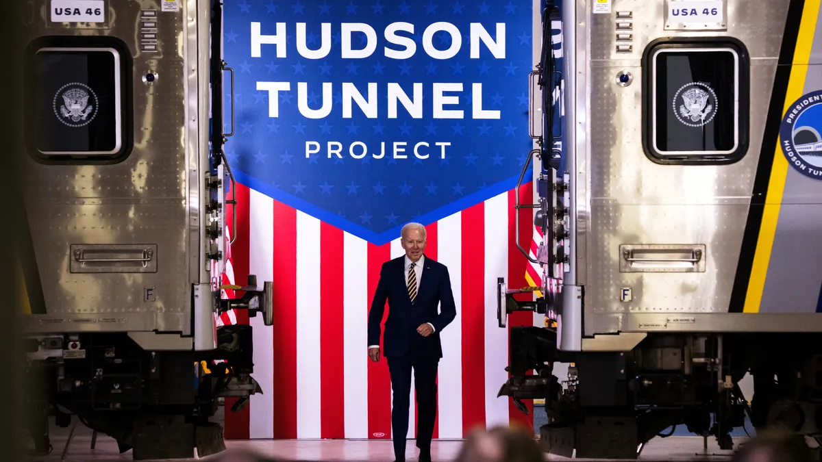 President Joe Biden dressed in a dark suit and striped tie walks onto a stage between two rail cars, with an american flag background that says "Hudson Tunnel project."