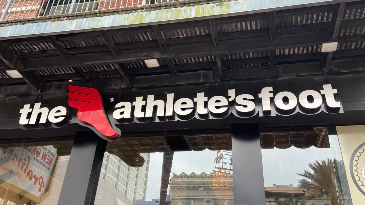 Buildings and signs are reflected in a store window. Above a sign in white letters reads "the athlete's foot," with a red symbol of a winged foot.