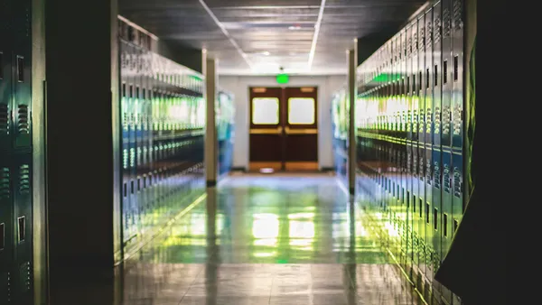 An empty school hallway looks straight at two doors. Over the doors is an exit sign. There is a green glow from the exit sign.