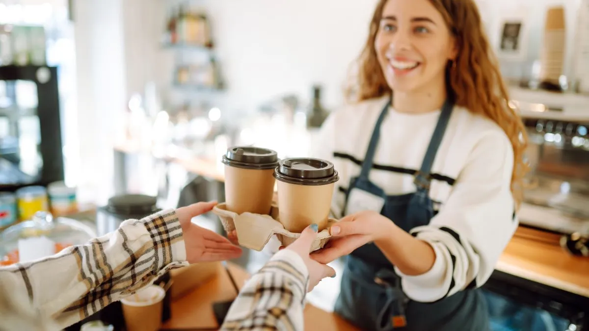 A smiling female barista hands two warm takeout coffee cups to a waiting customer.