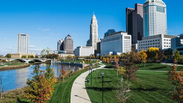 A daytime shot of Columbus, Ohio, with a bridge, river and green space in the foreground. Tall buildings dot the background.