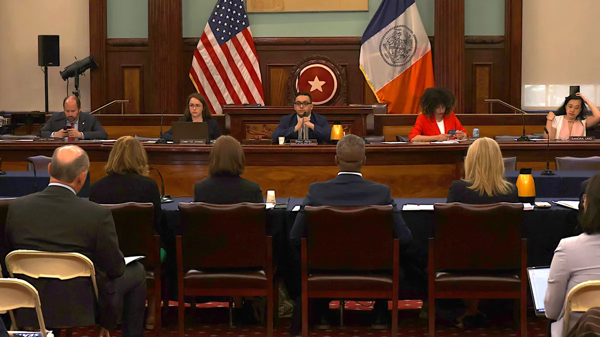 Several people sit at a raised wooden desk behind a U.S. and New York state flag. Facing them are another row of people dressed in suits.