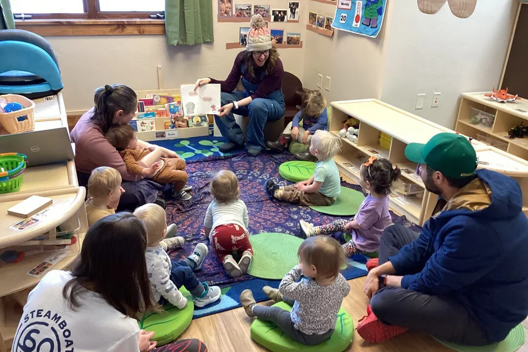 A woman in ski gear sits on a chair reading to a group of children in a day care classroom.