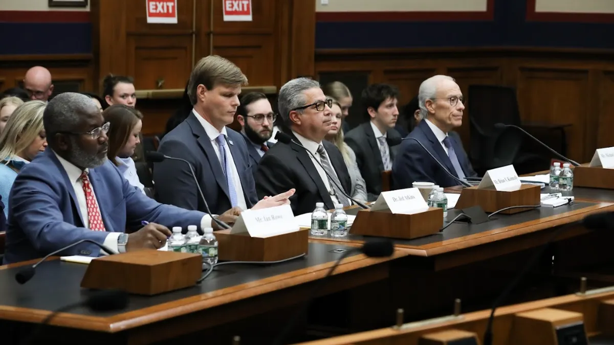 Four people sit at a table. On the table are microphones. People seated in rows are behind them.