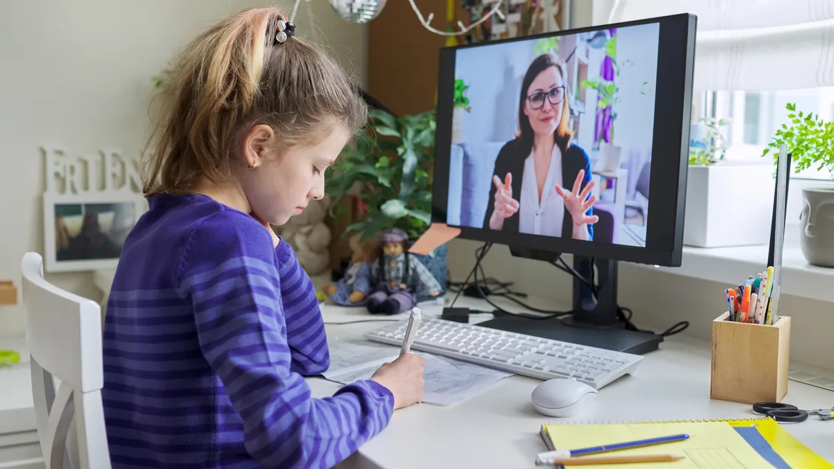 A student sits at a desk, holding a pen and looking down at a paper. In front of the student is a computer monitor and the screen shows an adult holding up fingers.