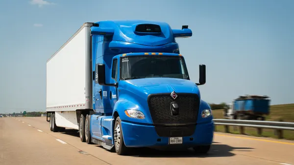 A blue tractor equipped with a white trailer and self-driving technology drives on a road.