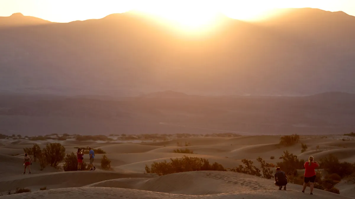 People watch a hazy sun rise while standing on desert dunes.