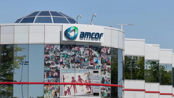 A modern white commercial building with reflective glass windows features a prominent "Amcor" sign at the top. The front of the building displays a collage of employee and family photos, along with a large safety sign that reads, "We work safely because our families are waiting for us at home." 