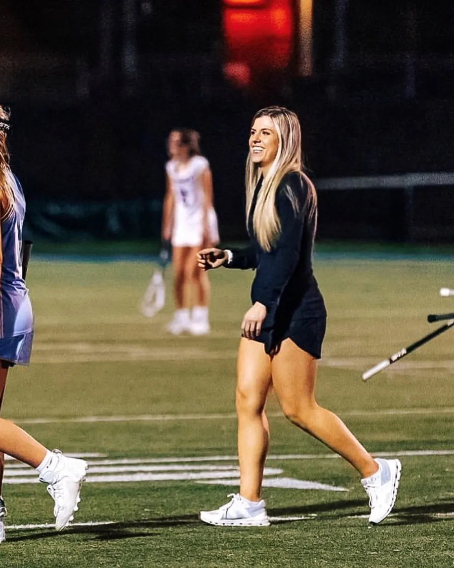 An adult is walking across a game field. A few lacrosse players are seen in the background